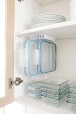 Clear glass food storage containers with light blue silicone seals hanging on a white kitchen cabinet shelf, alongside stacked white plates and more glass containers.